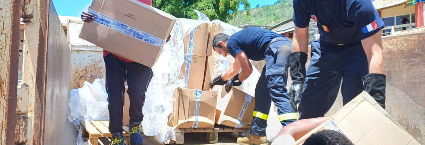 Texte alt en français : Des volontaires et des membres des services de secours français déchargent un camion rempli de cartons d'aide humanitaire sous un ciel bleu. Certains portent des uniformes avec des écussons tricolores, tandis que d'autres sont en tenue civile. Le groupe travaille ensemble pour distribuer des fournitures essentielles, illustrant un effort solidaire et humanitaire.  Texte alt en anglais : Volunteers and members of the French emergency services unload a truck filled with humanitarian ai