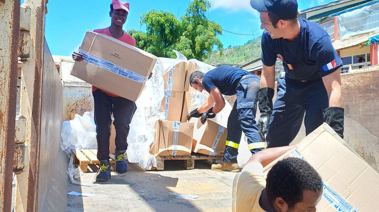 Texte alt en français : Des volontaires et des membres des services de secours français déchargent un camion rempli de cartons d'aide humanitaire sous un ciel bleu. Certains portent des uniformes avec des écussons tricolores, tandis que d'autres sont en tenue civile. Le groupe travaille ensemble pour distribuer des fournitures essentielles, illustrant un effort solidaire et humanitaire.  Texte alt en anglais : Volunteers and members of the French emergency services unload a truck filled with humanitarian ai