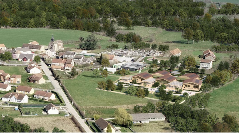 Vue aérienne d’un village entouré de verdure avec un nouveau complexe scolaire en bois intégré dans le paysage rural. L’école, composée de plusieurs bâtiments à toits inclinés, est entourée d’arbres et d’espaces verts. À proximité, on distingue des maisons, une église et un cimetière, illustrant le cadre authentique et paisible du village. L’architecture moderne de l’école s’harmonise avec l’environnement naturel et traditionnel.