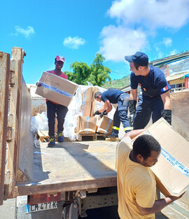 Texte alt en français : Des volontaires et des membres des services de secours français déchargent un camion rempli de cartons d'aide humanitaire sous un ciel bleu. Certains portent des uniformes avec des écussons tricolores, tandis que d'autres sont en tenue civile. Le groupe travaille ensemble pour distribuer des fournitures essentielles, illustrant un effort solidaire et humanitaire.  Texte alt en anglais : Volunteers and members of the French emergency services unload a truck filled with humanitarian ai