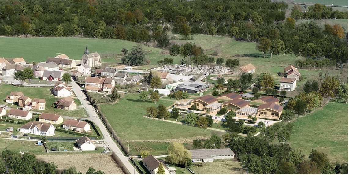 Vue aérienne d’un village entouré de verdure avec un nouveau complexe scolaire en bois intégré dans le paysage rural. L’école, composée de plusieurs bâtiments à toits inclinés, est entourée d’arbres et d’espaces verts. À proximité, on distingue des maisons, une église et un cimetière, illustrant le cadre authentique et paisible du village. L’architecture moderne de l’école s’harmonise avec l’environnement naturel et traditionnel.