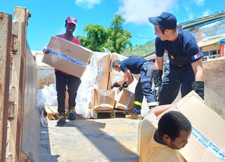 Texte alt en français : Des volontaires et des membres des services de secours français déchargent un camion rempli de cartons d'aide humanitaire sous un ciel bleu. Certains portent des uniformes avec des écussons tricolores, tandis que d'autres sont en tenue civile. Le groupe travaille ensemble pour distribuer des fournitures essentielles, illustrant un effort solidaire et humanitaire.  Texte alt en anglais : Volunteers and members of the French emergency services unload a truck filled with humanitarian ai